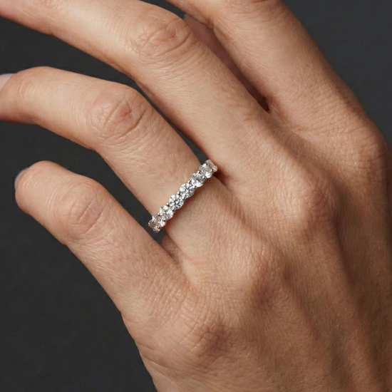 A model's hand wearing a delicate white gold or platinum half-eternity ring featuring round-cut diamonds against a dark background.