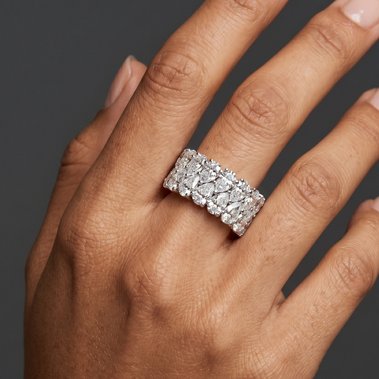 A close-up of an elegantly posed hand with medium-tan skin wearing a wide, luxurious ring entirely encrusted with two staggered rows of pear-shaped diamonds. The hand rests against a solid dark charcoal gray background under soft, diffused studio lighting.