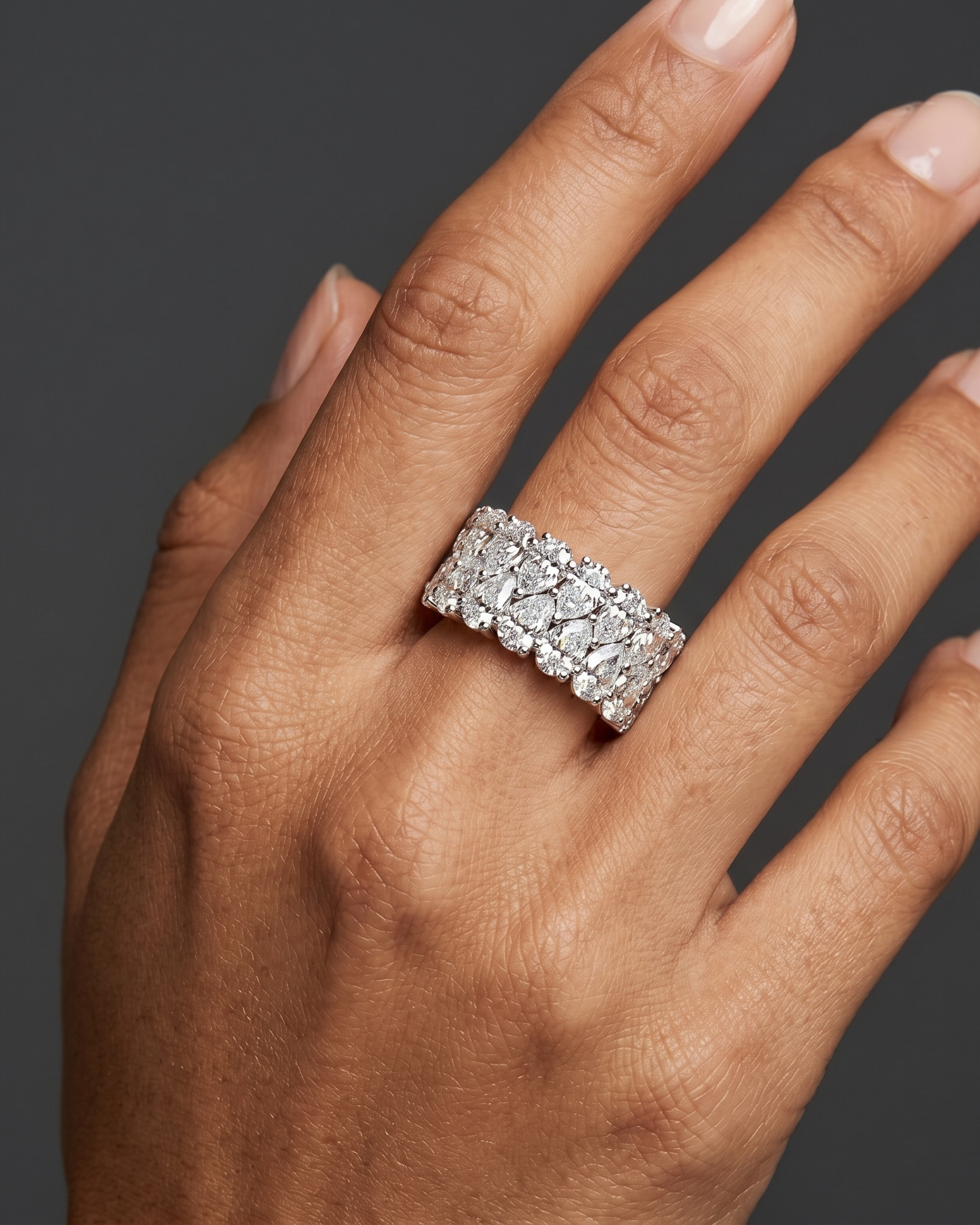 A close-up of an elegantly posed hand with medium-tan skin wearing a wide, luxurious ring entirely encrusted with two staggered rows of pear-shaped diamonds. The hand rests against a solid dark charcoal gray background under soft, diffused studio lighting.