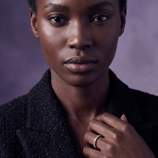 Gold diamond eternity band ring worn on the finger of a model in a black tweed blazer against a muted purple background.