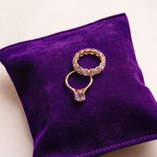 Yellow gold Amorati Bold Bezel 7-Carat Round Diamond Eternity Band displayed on a vibrant purple velvet pillow, styled with a round solitaire engagement ring.