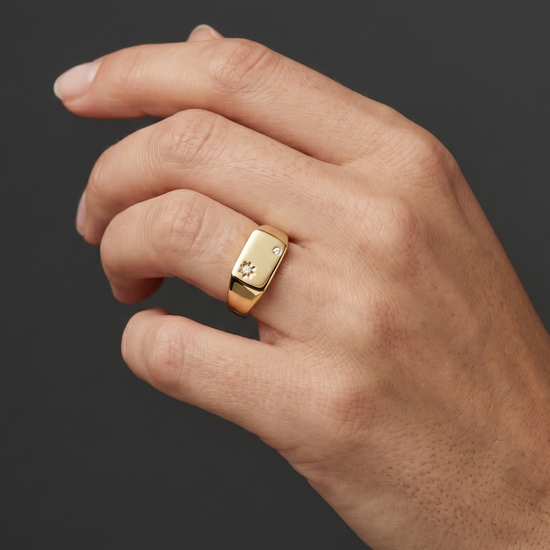 A close-up of a woman's hand wearing a petite, rectangular gold signet ring featuring two small diamonds, set against a dark charcoal gray background.