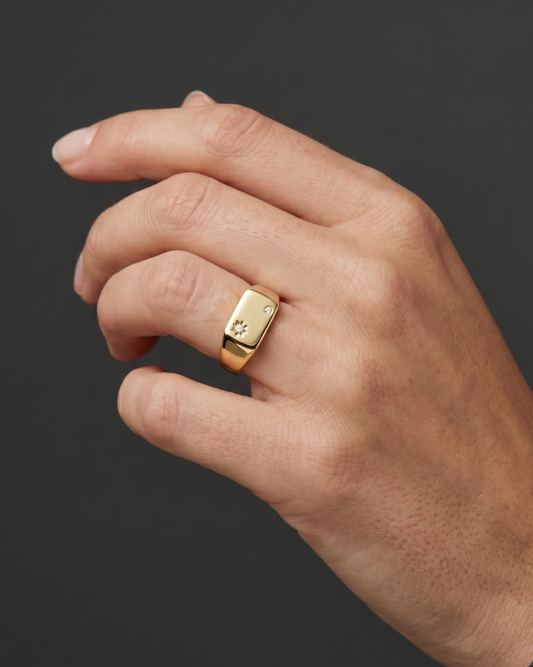 A close-up of a woman's hand wearing a petite, rectangular gold signet ring featuring two small diamonds, set against a dark charcoal gray background.