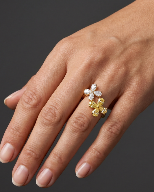 Close-up of a hand wearing a gold ring featuring two diamond flowers, one made of white diamonds and the other of yellow diamonds, against a dark gray background.