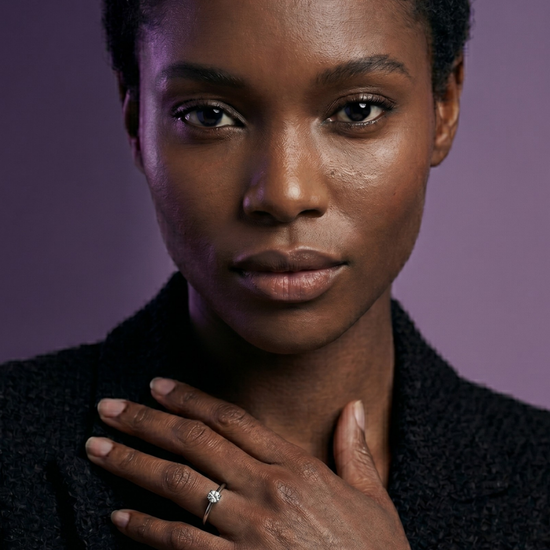Close-up portrait of a Black woman displaying a round brilliant-cut solitaire diamond ring on her left hand. She is wearing a textured black blazer against a moody purple background.
