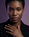 Close-up portrait of a Black woman displaying a round brilliant-cut solitaire diamond ring on her left hand. She is wearing a textured black blazer against a moody purple background.