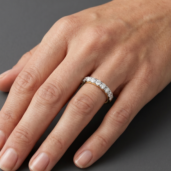 A close-up of a woman's hand resting elegantly on a dark charcoal gray background, wearing a petite gold ring with a row of small diamonds. Soft lighting highlights natural skin texture and the jewelry.