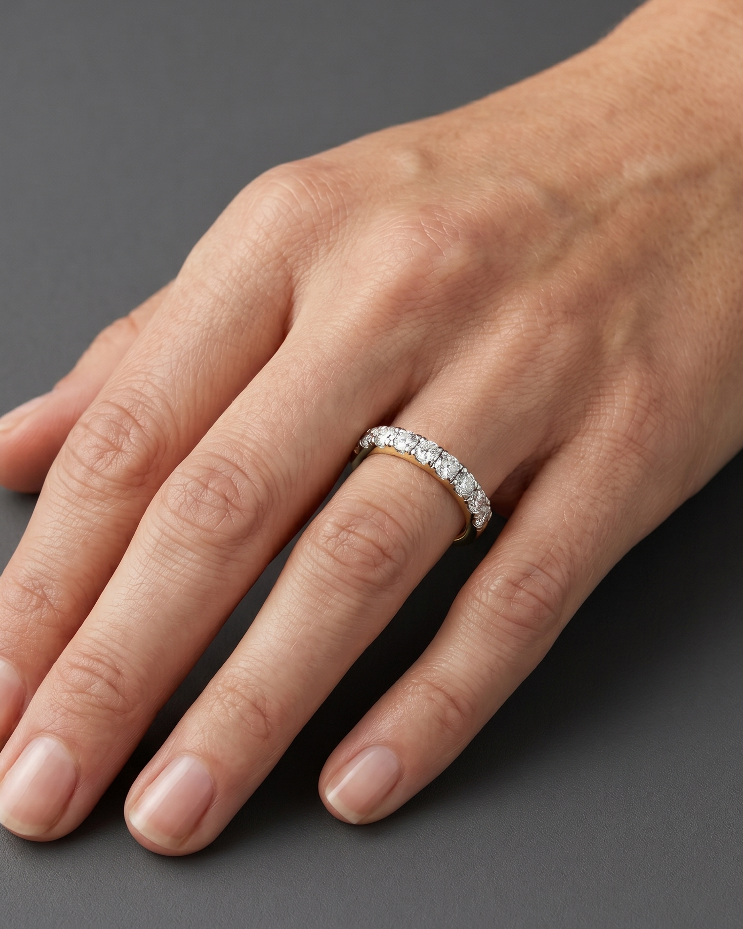 A close-up of a woman's hand resting elegantly on a dark charcoal gray background, wearing a petite gold ring with a row of small diamonds. Soft lighting highlights natural skin texture and the jewelry.