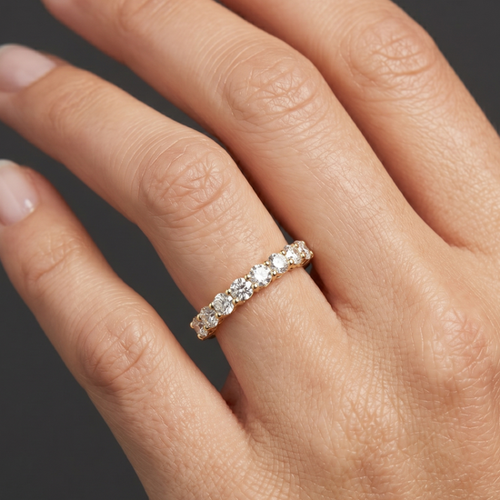 An editorial, close-up photograph focuses on a woman's hand, capturing her fingers and wrist against a smooth, dark charcoal gray background. She is wearing a petite, elegant gold ring set with a row of ten round, brilliant-cut diamonds that sparkle under soft, diffused studio lighting. The ring fits naturally on her ring finger, creating realistic skin compression, shadows, and subtle reflections on the polished metal. Her hand is posed gracefully, showcasing natural skin texture and well-manicured nails, 