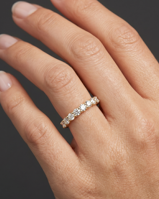 An editorial, close-up photograph focuses on a woman's hand, capturing her fingers and wrist against a smooth, dark charcoal gray background. She is wearing a petite, elegant gold ring set with a row of ten round, brilliant-cut diamonds that sparkle under soft, diffused studio lighting. The ring fits naturally on her ring finger, creating realistic skin compression, shadows, and subtle reflections on the polished metal. Her hand is posed gracefully, showcasing natural skin texture and well-manicured nails, 