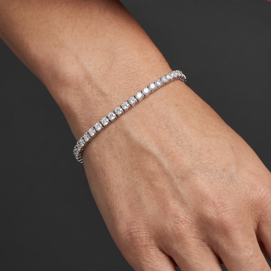 A close-up of a person's wrist and hand wearing a thin silver tennis bracelet with clear stones, set against a dark gray background.