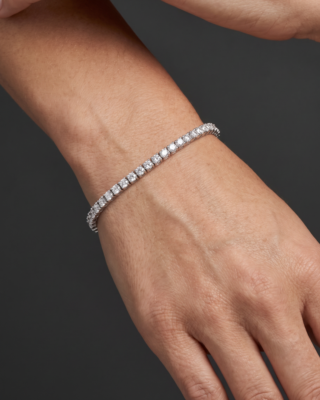 A close-up of a person's wrist and hand wearing a thin silver tennis bracelet with clear stones, set against a dark gray background.