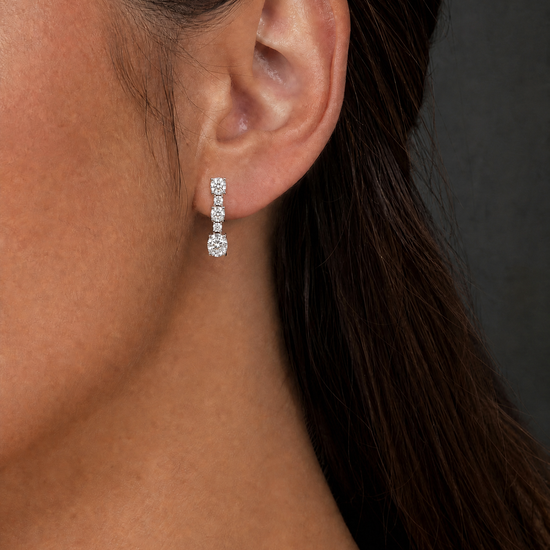 A close-up of a model's ear wearing a small silver drop earring with three clear round-cut stones, set against a dark gray background.