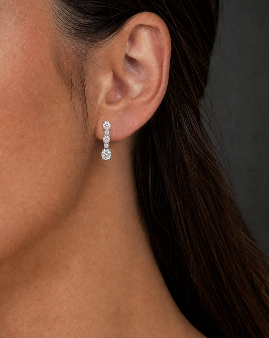 A close-up of a model's ear wearing a small silver drop earring with three clear round-cut stones, set against a dark gray background.