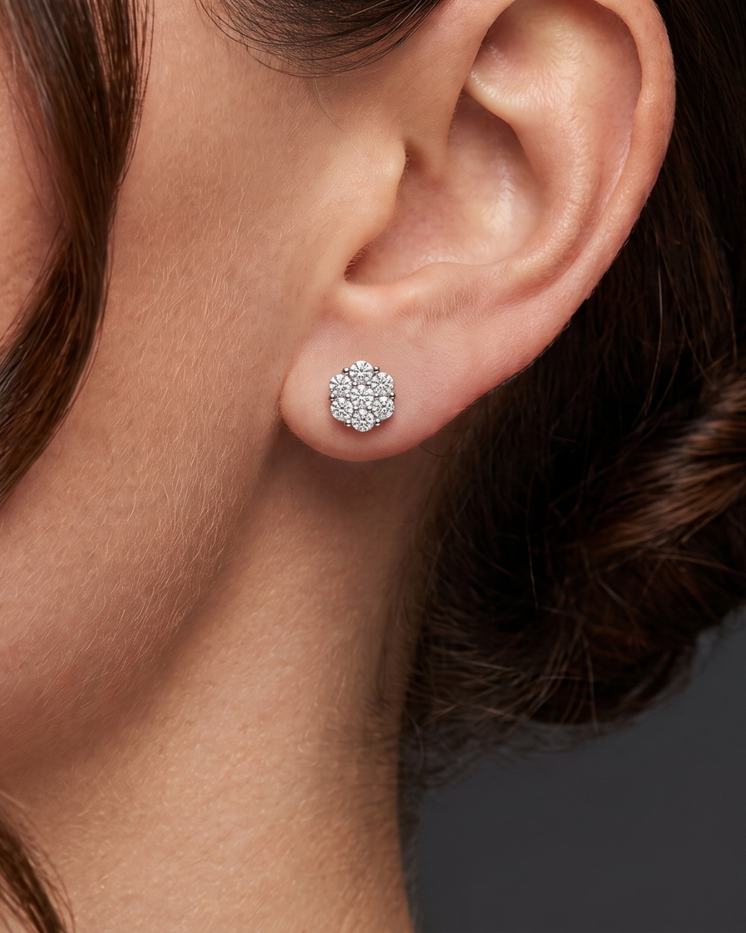 A close-up of a model's ear wearing a small, round cluster stud earring set with clear gemstones in a silver-toned metal.