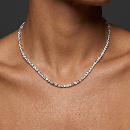 A close-up of a model's neck and upper chest wearing a delicate silver tennis necklace with small clear stones.