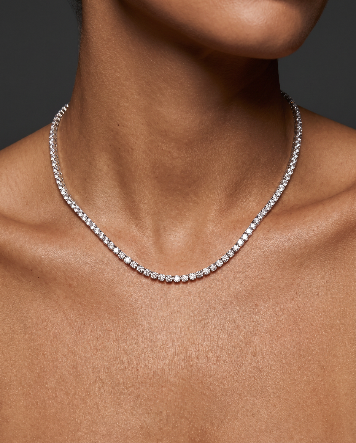 A close-up of a model's neck and upper chest wearing a delicate silver tennis necklace with small clear stones.
