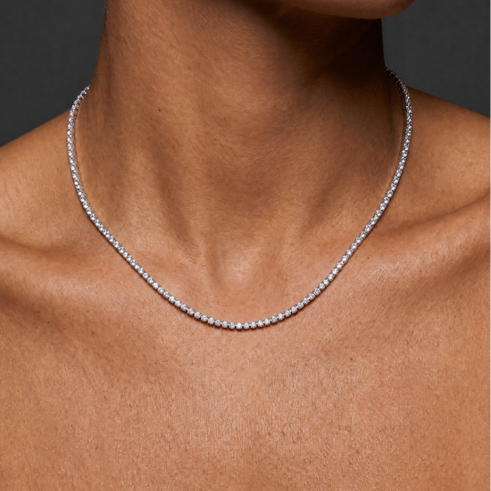 A close-up of a model's neck and upper chest wearing a delicate silver tennis necklace with small clear stones.