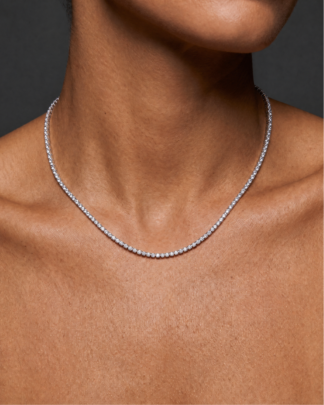 A close-up of a model's neck and upper chest wearing a delicate silver tennis necklace with small clear stones.