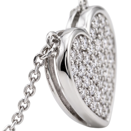 A close-up of a silver heart-shaped pendant necklace, partially open to reveal a heart-shaped locket filled with pavé-set clear gemstones, attached to a silver chain against a white background.