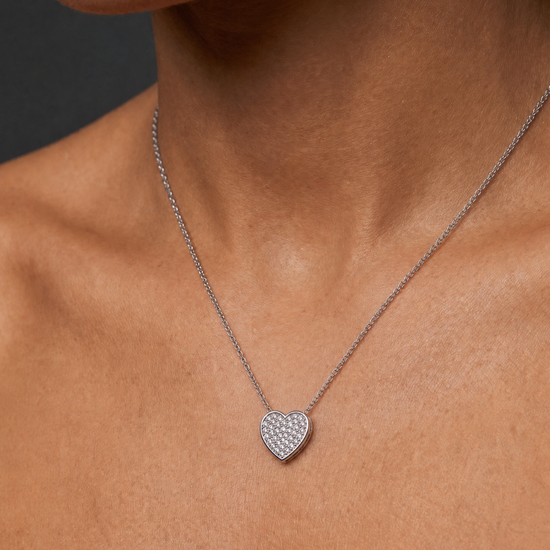 A close-up of a model's neck and chest wearing a delicate silver chain necklace with a small, heart-shaped pavé pendant.
