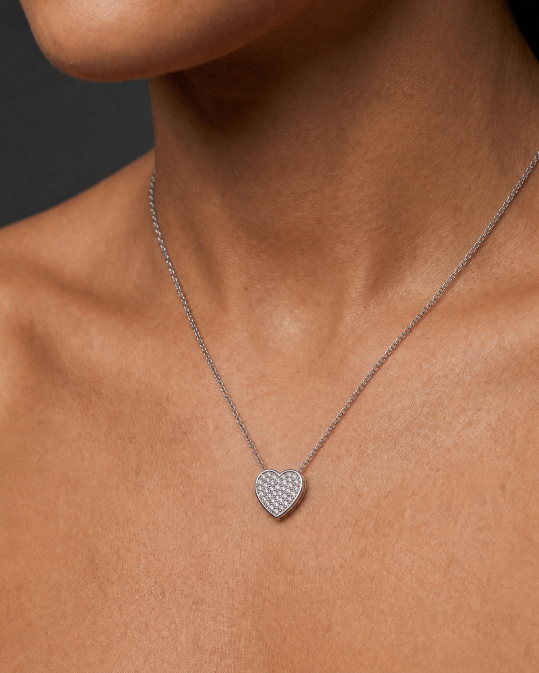 A close-up of a model's neck and chest wearing a delicate silver chain necklace with a small, heart-shaped pavé pendant.