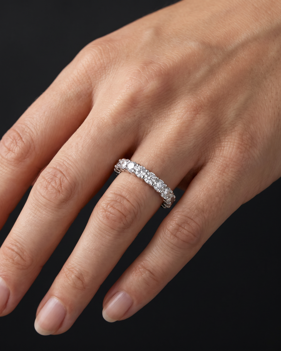 A close-up of a hand wearing a silver diamond eternity ring against a solid black background.