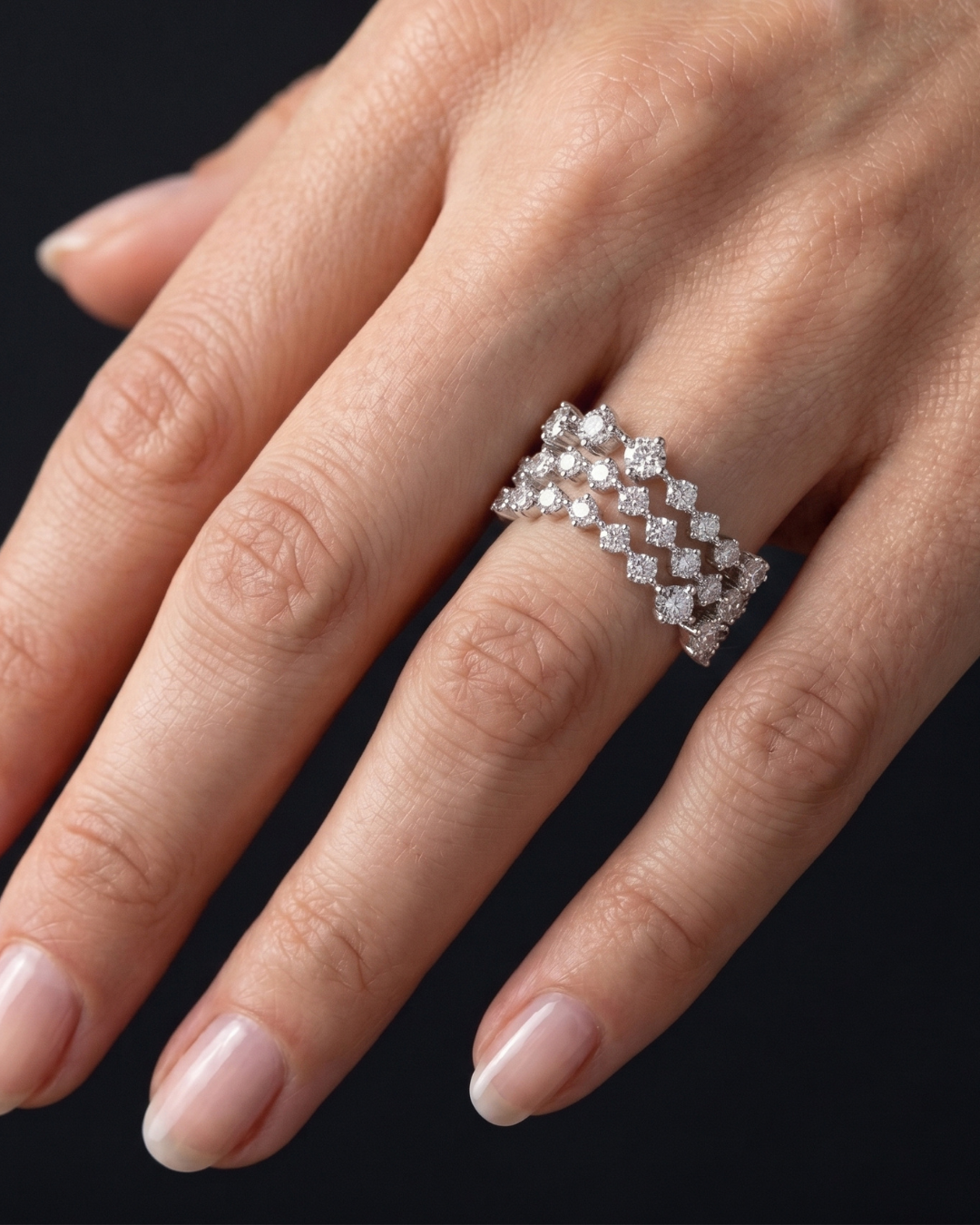 A close-up of a hand wearing a silver-toned diamond ring with a repeating geometric diamond pattern, set against a solid black background.