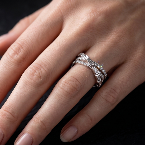 A close-up of a hand wearing three silver rings, including a diamond band and two stacked eternity-style bands, against a dark background.