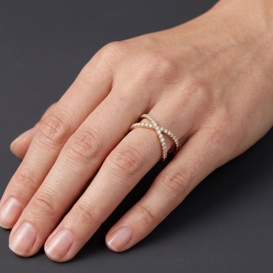 A close-up of a hand wearing a delicate double-band ring with small white stones set in a yellow gold-tone band, shown against a dark gray background.