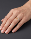 A close-up of a hand wearing a delicate double-band ring with small white stones set in a yellow gold-tone band, shown against a dark gray background.