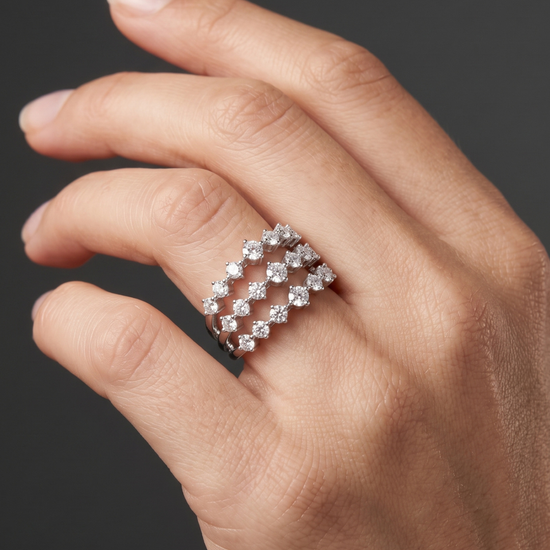 A close-up of a hand wearing a silver-toned ring with a double row of sparkling clear gemstones in a diamond-like pattern, set against a dark gray background.