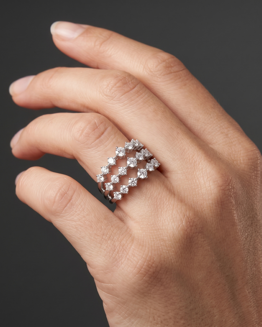 A close-up of a hand wearing a silver-toned ring with a double row of sparkling clear gemstones in a diamond-like pattern, set against a dark gray background.