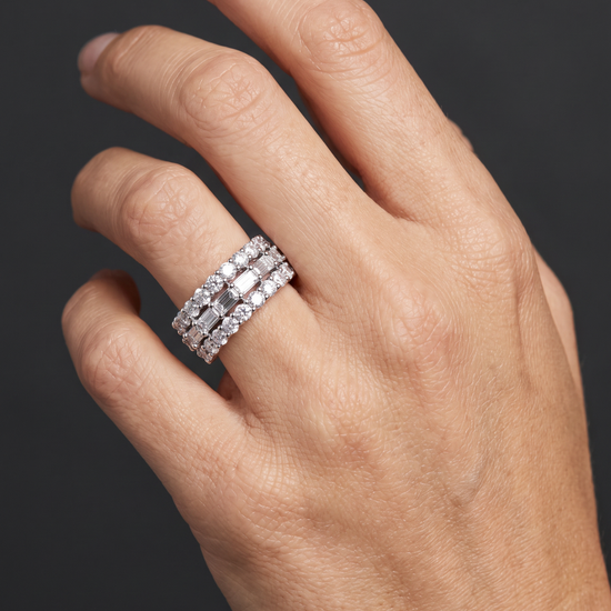 A close-up of a hand wearing a three-row diamond eternity ring with a silver-toned band, set against a dark gray background.
