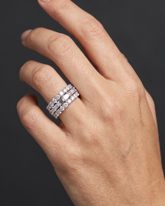 A close-up of a hand wearing a three-row diamond eternity ring with a silver-toned band, set against a dark gray background.