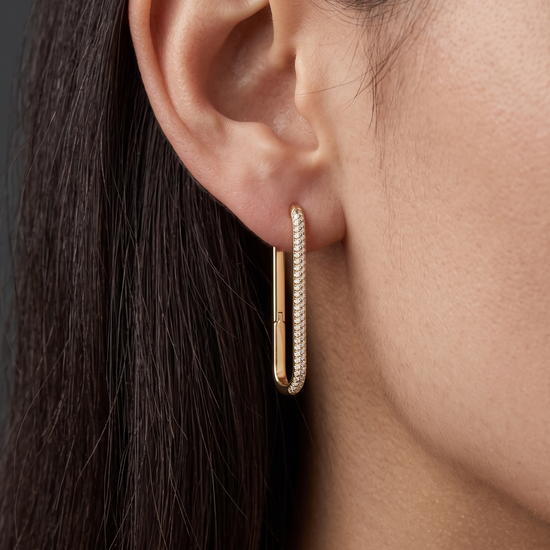 Close-up of a woman's ear wearing a diamond-lined gold rectangular hoop earring. Dark gray background.