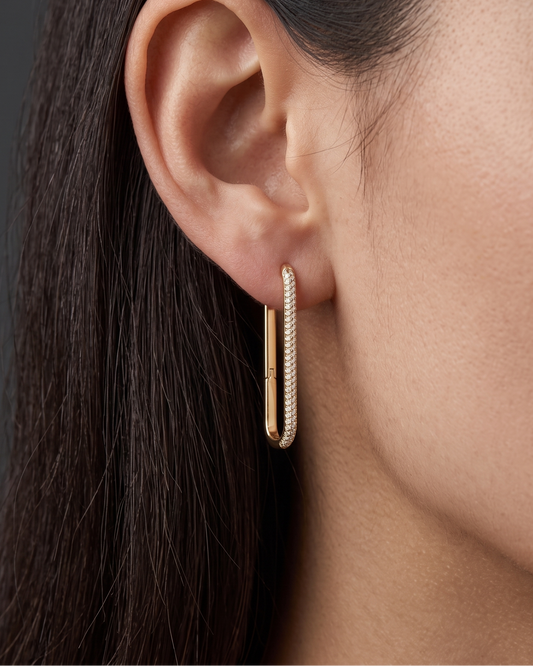 Close-up of a woman's ear wearing a diamond-lined gold rectangular hoop earring. Dark gray background.