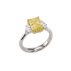 A three-quarter angled view of a three-stone engagement ring on a black background. The ring features a light, pastel yellow emerald-cut center diamond secured by yellow gold prongs, with clear white diamonds set on either side. The ring has a rounded, polished white metal band.