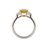 A profile view of an engagement ring showing the full circular white metal band and the side of the setting against a black background. The center stone is a pastel yellow emerald-cut diamond resting in an elevated yellow gold basket setting with an open gallery. Clear white side diamonds are visible sloping down the shoulders of the band.
