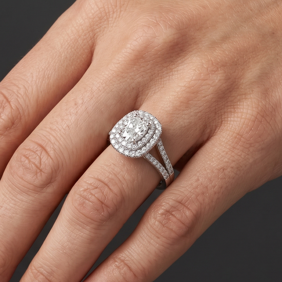 Close-up of a woman's elegantly posed hand wearing a sparkling diamond engagement ring. The ring features an elongated center diamond surrounded by a double halo of smaller diamonds and a diamond-encrusted split-shank band. The background is a smooth, dark charcoal gray.