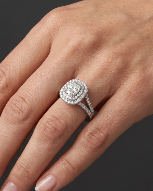 Close-up of a woman's elegantly posed hand wearing a sparkling diamond engagement ring. The ring features an elongated center diamond surrounded by a double halo of smaller diamonds and a diamond-encrusted split-shank band. The background is a smooth, dark charcoal gray.