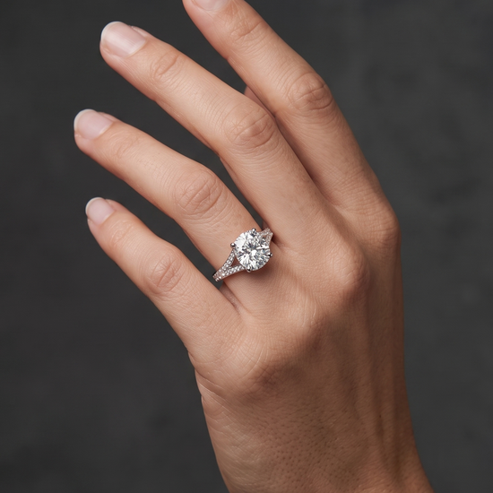 A close-up editorial photograph of a woman's elegantly posed hand against a smooth, dark charcoal gray background. She is wearing a luxurious silver-tone engagement ring featuring a large, brilliant elongated cushion-cut center diamond and a diamond-encrusted split-shank band.
