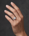 A close-up editorial photograph of a woman's elegantly posed hand against a smooth, dark charcoal gray background. She is wearing a luxurious silver-tone engagement ring featuring a large, brilliant elongated cushion-cut center diamond and a diamond-encrusted split-shank band.