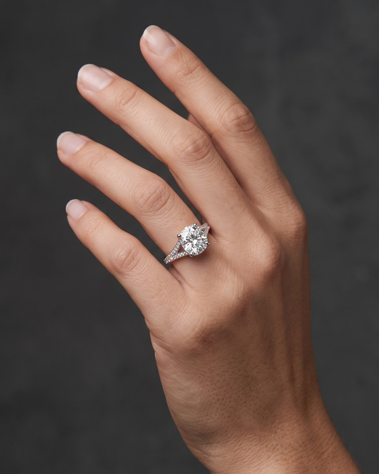 A close-up editorial photograph of a woman's elegantly posed hand against a smooth, dark charcoal gray background. She is wearing a luxurious silver-tone engagement ring featuring a large, brilliant elongated cushion-cut center diamond and a diamond-encrusted split-shank band.