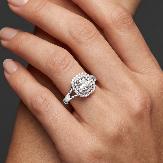 A close-up photograph of a woman's crossed hands against a dark gray background, featuring a sparkling double-halo diamond engagement ring with a split-shank white metal band.