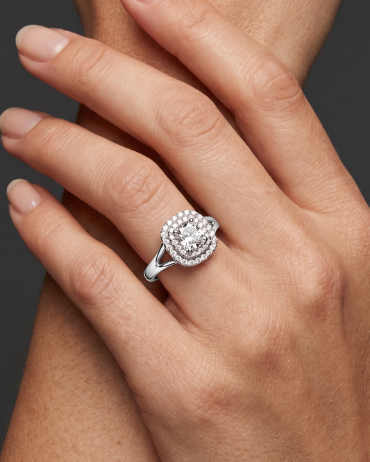 A close-up photograph of a woman's crossed hands against a dark gray background, featuring a sparkling double-halo diamond engagement ring with a split-shank white metal band.