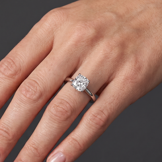 Close-up of a woman's hand resting elegantly against a smooth, dark charcoal gray background, wearing a silver engagement ring. The ring features a modest-sized, elongated cushion-cut diamond set on a split-shank band. Natural lighting highlights the sparkle of the gemstone and the realistic texture of the skin.