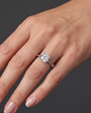 Close-up of a woman's hand resting elegantly against a smooth, dark charcoal gray background, wearing a silver engagement ring. The ring features a modest-sized, elongated cushion-cut diamond set on a split-shank band. Natural lighting highlights the sparkle of the gemstone and the realistic texture of the skin.