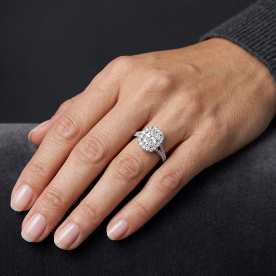 A close-up photograph of a woman's hand resting gracefully on a plush, dark gray surface, showcasing a sparkling diamond engagement ring on her ring finger. The ring features an elongated cushion-cut center diamond surrounded by a diamond halo, set on a split-shank band encrusted with smaller diamonds. Soft, diffused lighting highlights the brilliance of the ring and the natural texture of the skin against a smooth, dark charcoal gray background.