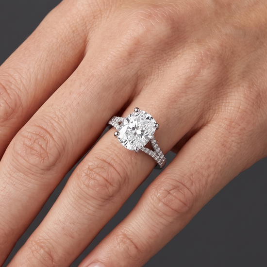Close-up of a woman's hand elegantly posed against a dark charcoal gray background, wearing a white gold or platinum engagement ring with a large cushion-cut diamond and a sparkling split-shank band.